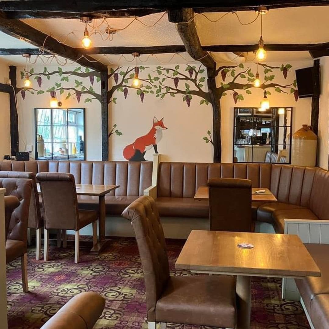 Fox and Grapes Pudsey cozy interior fox mural wooden beams Interior of Fox & Grapes – Pudsey, showing a cozy dining area with exposed wooden beams, leather seating, wooden tables, and a whimsical wall mural featuring a fox and grapevines.