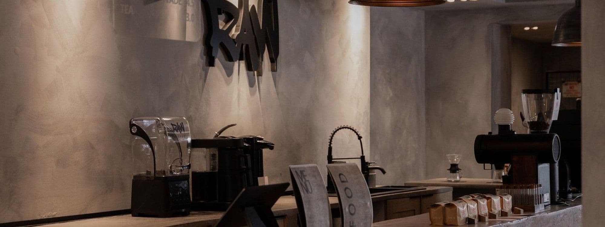 Interior view of Raw Coffee shop with espresso machines and coffee grinders on the counter, large 'RAW' sign on the wall, and coffee accessories on display.
