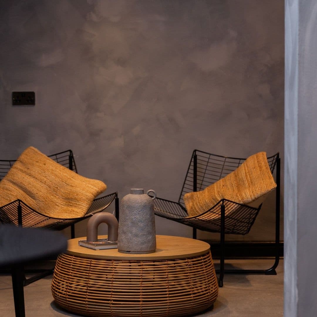 RawCoffee_ModernMinimalistInterior_ComfortableSeating Interior of Raw Coffee featuring modern minimalist furniture with a round wicker coffee table, black metal chairs with tan cushions, and a textured grey wall.
