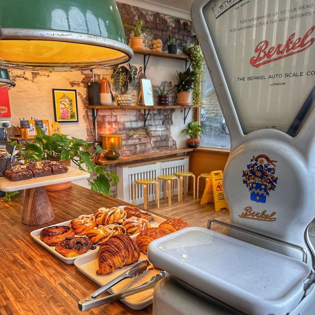 Silvers Deli Cosy Interior Fresh Pastries Vintage Scale Interior of Silver's Deli featuring a cozy setting with exposed brick walls, green plants, and vintage décor. A selection of freshly baked pastries, including chocolate cake, croissants, and cinnamon swirls, displayed on a wooden counter beside a classic Berkel weighing scale.