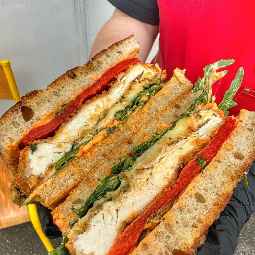 Silvers Deli Gourmet Chicken Sandwich Fresh Vegetables Close-up of a freshly made sandwich from Silver’s Deli, featuring sliced chicken, roasted red peppers, melted cheese, and greens between toasted bread slices, held by a person in a red shirt.