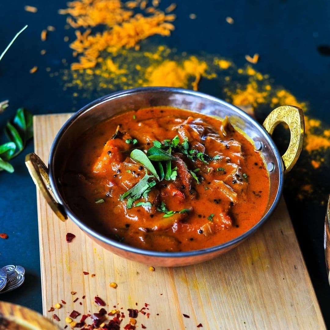 The Cats Pyjamas Chapel Allerton Delicious Curry Dish Aromatic Indian curry served in a traditional metal bowl at The Cat’s Pyjamas – Chapel Allerton, garnished with fresh herbs on a wooden board with vibrant spices scattered in the background.