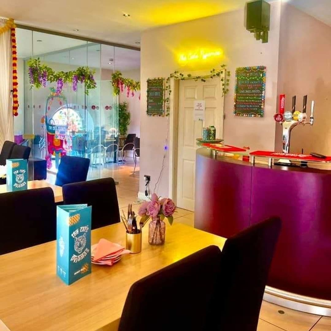 The Cats Pyjamas Farsley Colorful Interior Bar Area Decor Interior of The Cat’s Pyjamas – Farsley, showing a vibrant bar area with a purple counter, beer taps, and a menu board. The dining space is adorned with hanging plants, colorful decorations, and has a warm, inviting atmosphere with natural light.