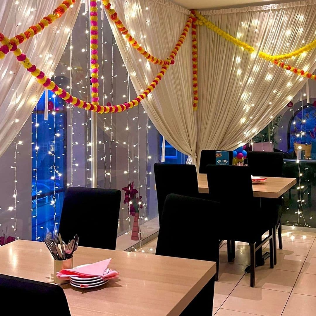The Cats Pyjamas Farsley Festive Lights Dining Ambiance Interior of The Cat’s Pyjamas – Farsley, showing a warmly lit dining area with draped ceiling fabric, fairy lights, and festive garland decorations. Wooden tables with black chairs and neatly arranged tableware are visible.