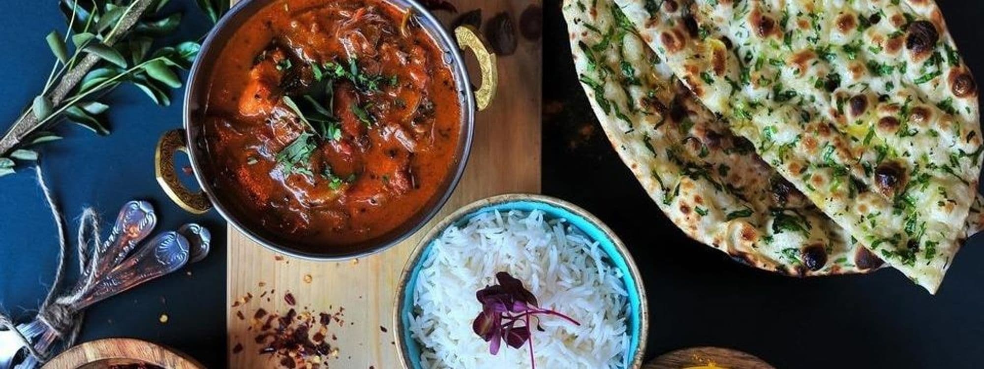 Delicious Indian cuisine served at The Cat’s Pyjamas – Farsley, featuring a bowl of rich, red curry garnished with herbs, a bowl of steamed white rice, and a golden-brown garlic naan bread.
