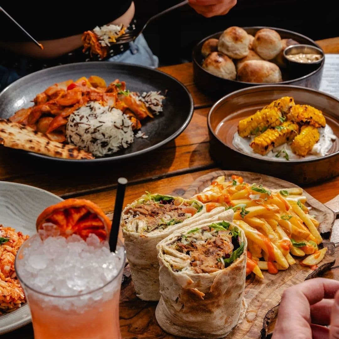 Treehouse Bar and Kitchen Delicious Food Spread Cocktail Assorted dishes and a cocktail on a wooden table at Treehouse Bar and Kitchen, showcasing a variety of meals including a wrap with fries, grilled corn, a bowl of curry with rice and naan, and bread rolls.