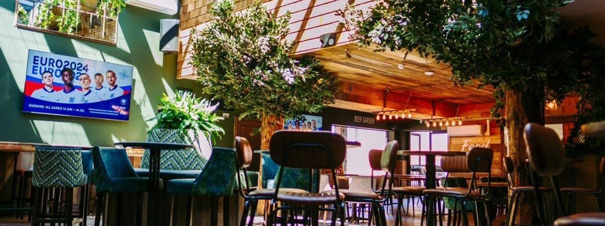 Interior of Treehouse Bar and Kitchen featuring a cozy and vibrant setting with plush seating, wooden tables, lush greenery, and a flat-screen TV displaying a sports event advertisement.