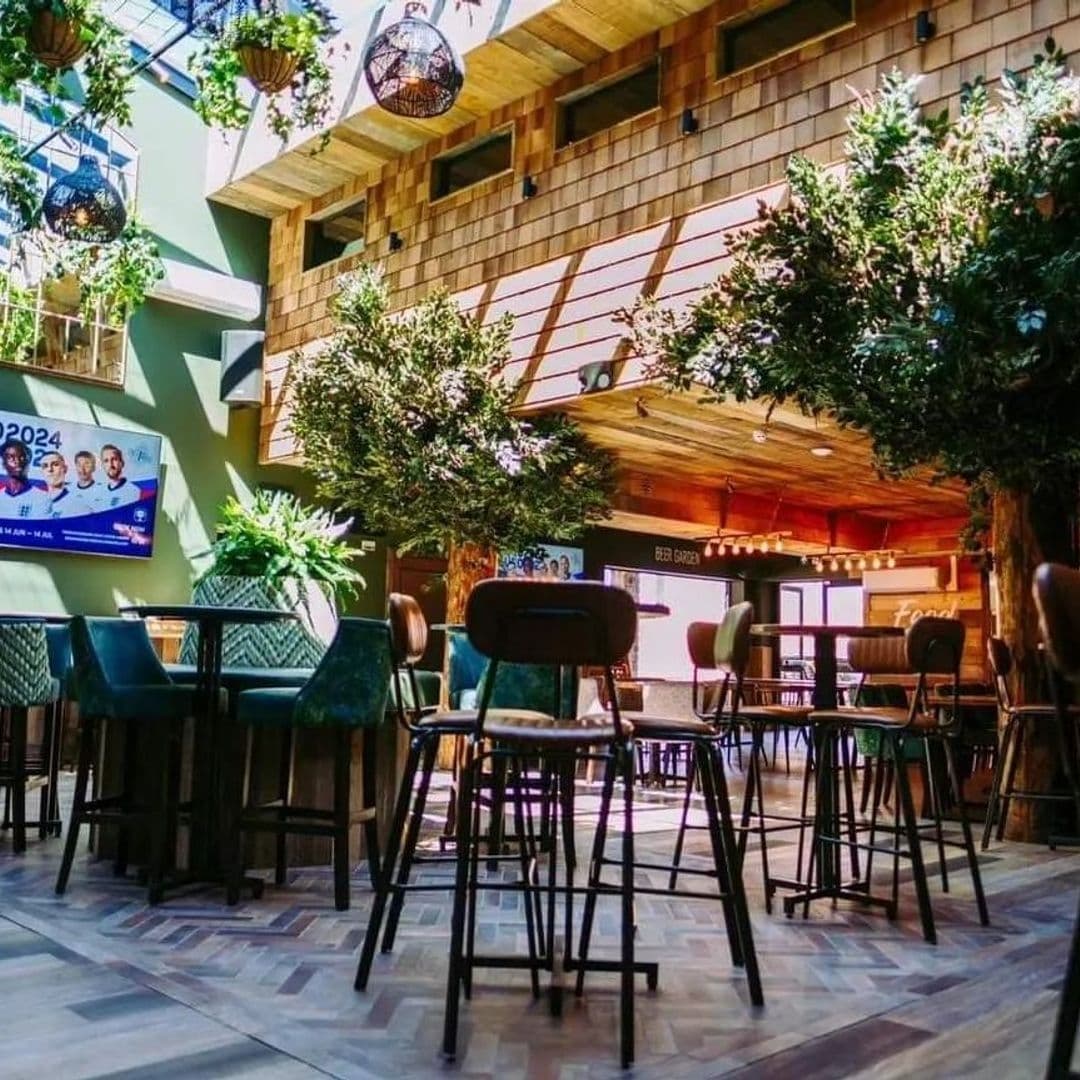 Treehouse_Bar_and_Kitchen_Sunny_Atrium_Style_Seating Interior of Treehouse Bar and Kitchen showing a cozy seating area with high stools, wooden tables, lush green plants, and a warm, inviting atmosphere under a high ceiling with hanging globe lights.