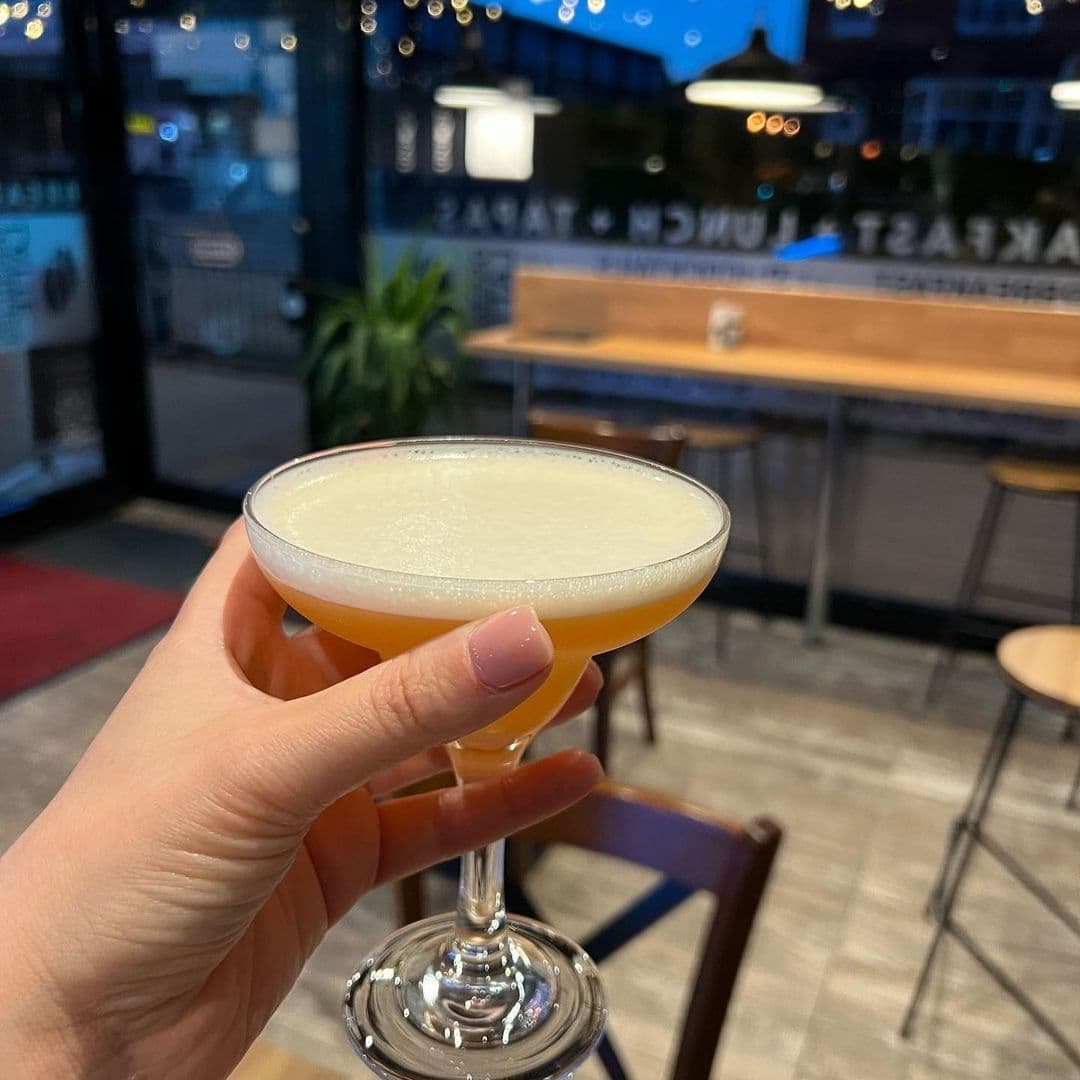 Unique Coffee Bistro Handheld Cocktail Evening Relaxation Hand holding a cocktail at Unique Coffee&Bistro with interior seating and window reflections in the background.