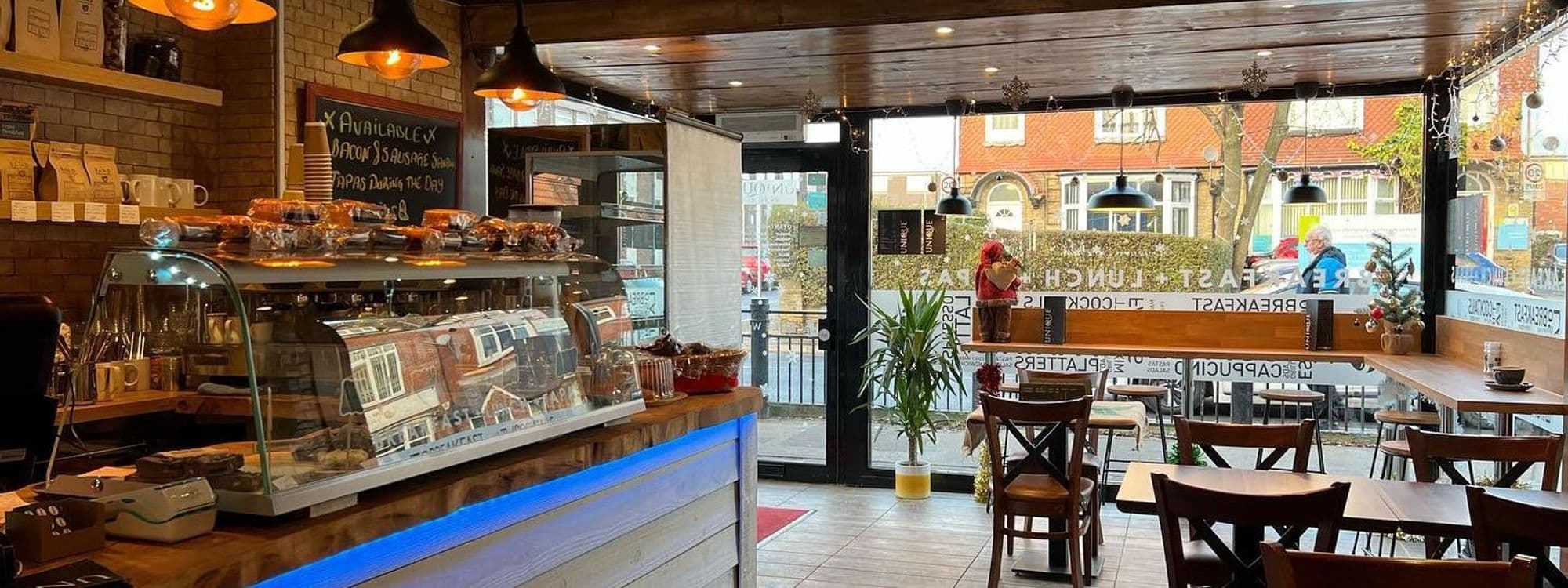 Interior of Unique Coffee&Bistro with wooden tables and chairs, a display case of pastries, and pendant lighting. A cozy atmosphere with a view of the street through large windows.