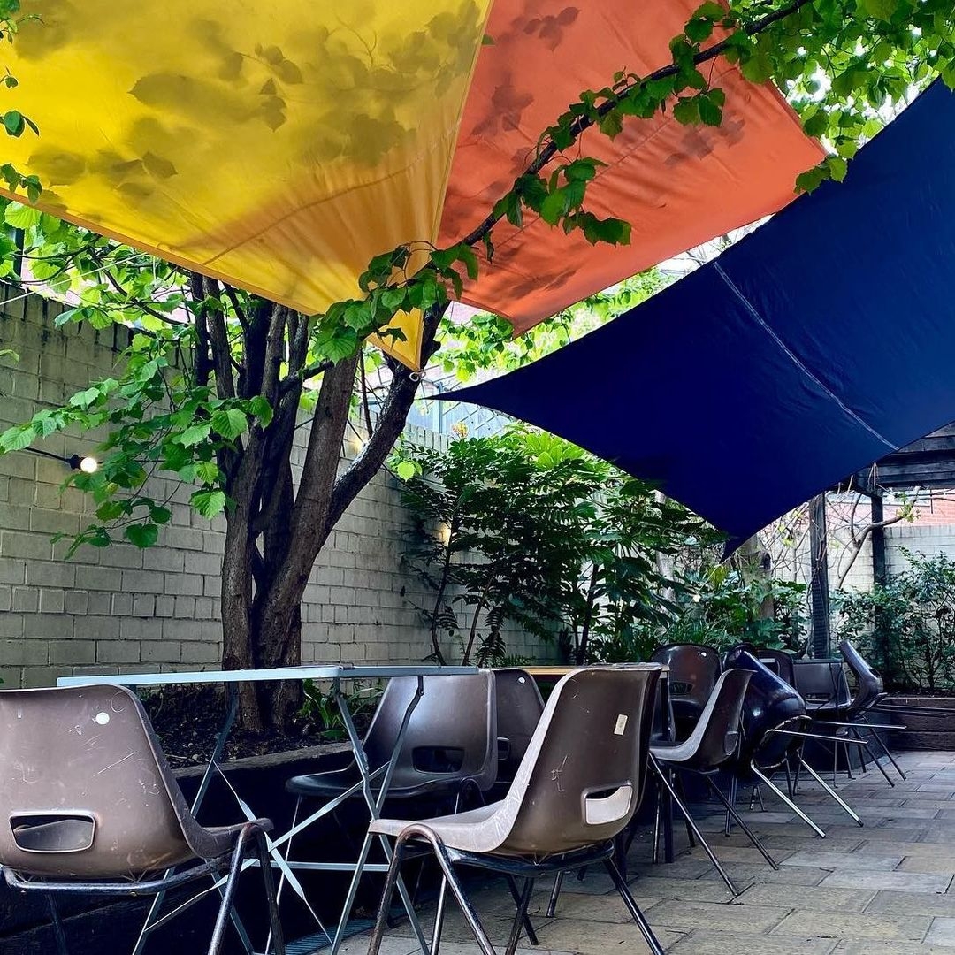 Wharf Chambers Outdoor Seating Colorful Canopies Relaxing Ambiance Outdoor seating area at Wharf Chambers with colorful overhead shades and a row of brown chairs against a backdrop of green foliage and a brick wall.