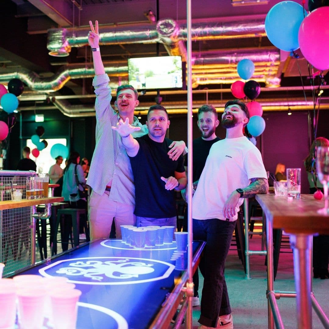 Boom Battle Bar Friends Enjoying Beer Pong Nightlife Group of friends enjoying a game of beer pong at Boom Battle Bar with colorful balloons and vibrant lighting in the background.