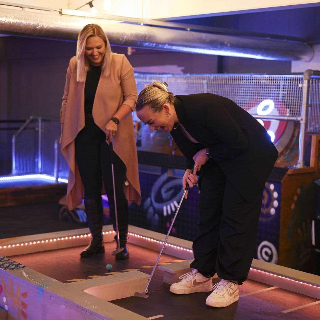 Boom Battle Bar Friends Playing Mini Golf Fun Evening Two women enjoying a game of mini-golf at Boom Battle Bar with neon lights in the background.