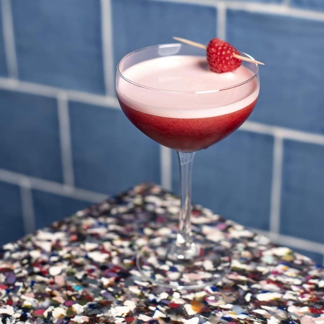 Electric_Shuffle_Signature_Cocktail_Raspberry_Garnish Craft cocktail with a frothy top and a raspberry garnish served at Electric Shuffle on a speckled countertop with a blue tile background.