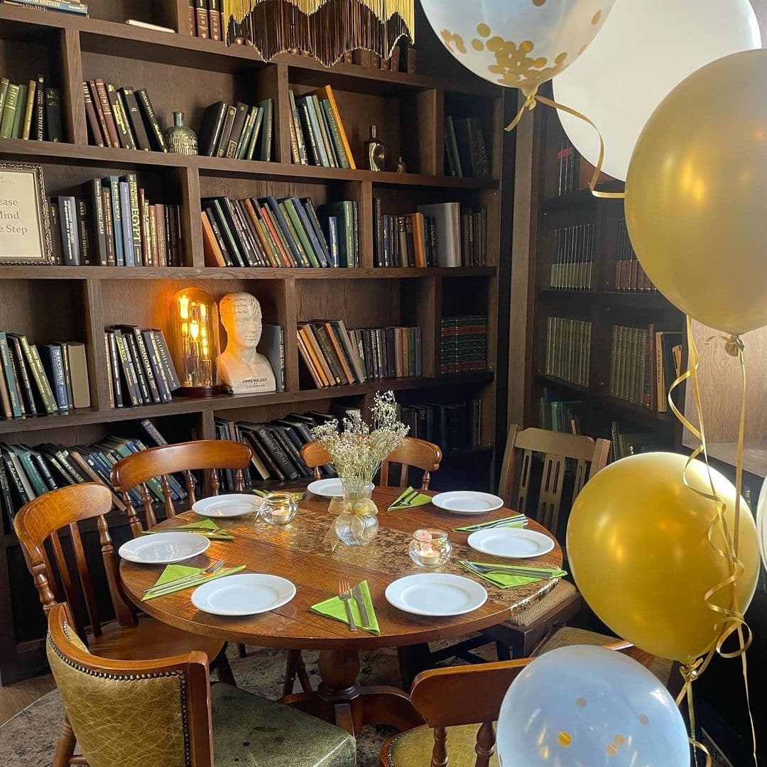 LambAndFlag_BookshelfDecor_ElegantDining_GoldenBalloons Elegant dining setup at Lamb And Flag with a wooden table set for a celebration, surrounded by classic chairs, golden balloons, and a bookshelf backdrop.