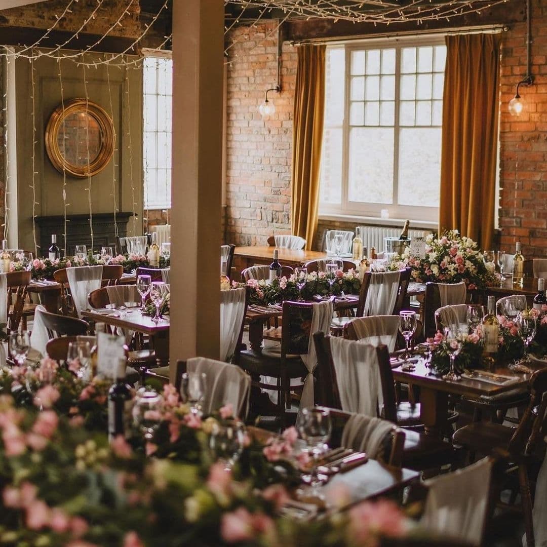 LambAndFlag_ElegantDiningSetup_BrickWalls_WarmLighting Elegant dining setup at Lamb And Flag with wooden tables adorned with floral centerpieces, white draped chairs, and twinkling string lights against exposed brick walls.