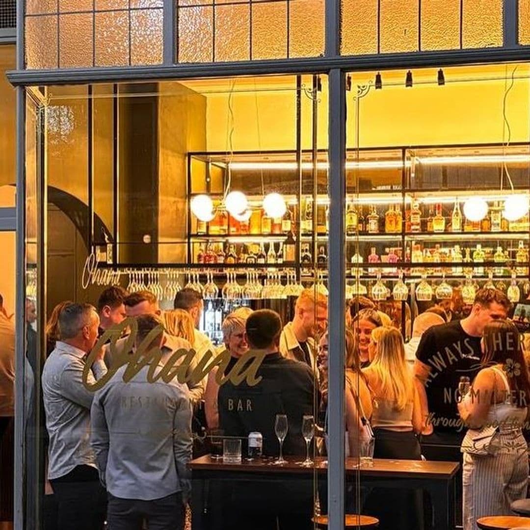 OHANA_vibrant bar scene social gathering people enjoying Vibrant atmosphere inside OHANA bar with patrons socializing and enjoying drinks, viewed through a glass window with the establishment's name prominently displayed.