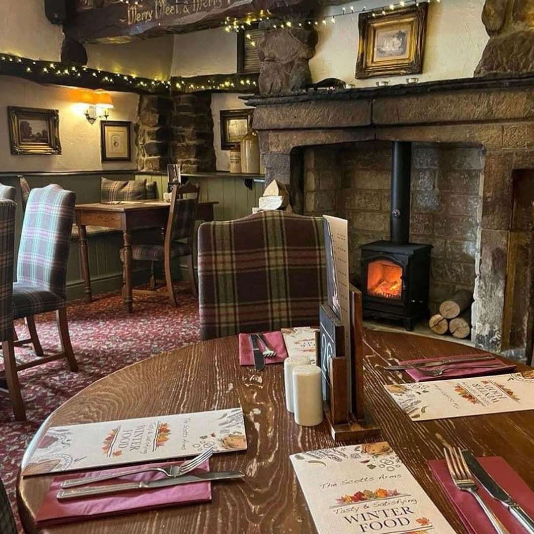 ScottsArms CozyPubInterior Fireplace WoodenFurniture Cozy interior of Scotts Arms with traditional wooden furniture, tartan upholstered chairs, a lit wood stove, and dining tables set with menus.
