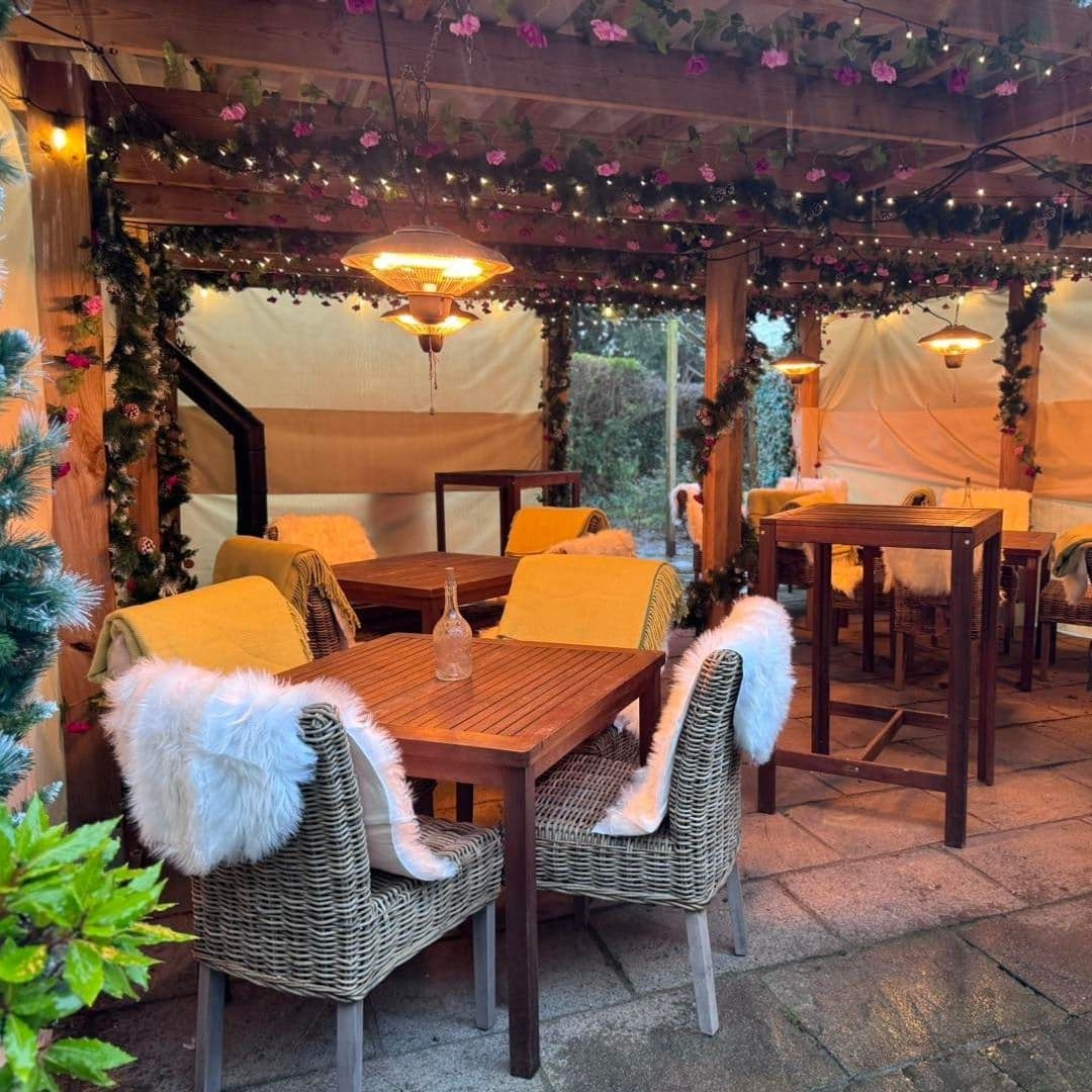 ScottsArms_CozyOutdoorSeating_FestiveDecor_WarmLighting Cozy outdoor seating area at Scotts Arms with wooden tables, wicker chairs with white fur, yellow cushions, and overhead floral decorations.