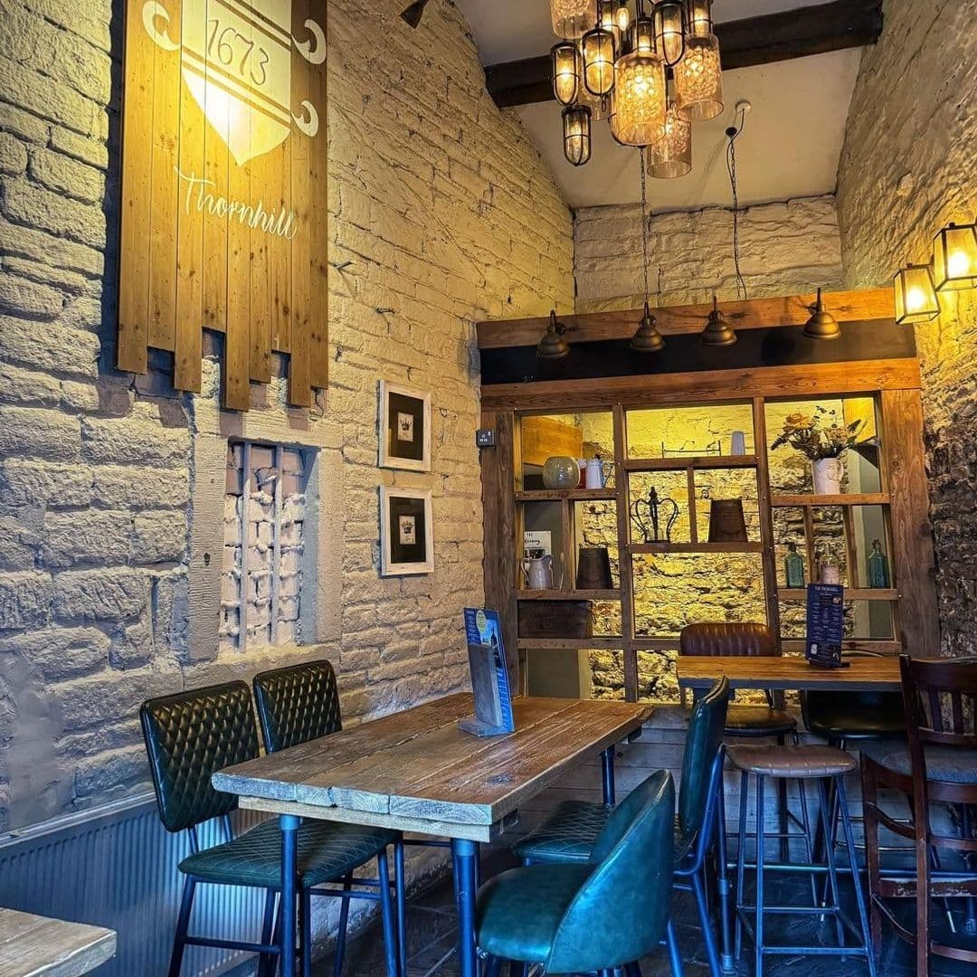 The Thornhill Cozy Interior Stone Walls Vintage Decor Cozy interior of The Thornhill featuring rustic stone walls, wooden tables with green velvet chairs, and warm lighting from unique hanging glass lanterns and wall-mounted lamps.