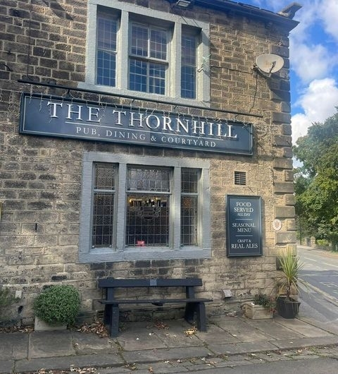 TheThornhill_HistoricPub_StoneBuilding_Bench_PlantPots Exterior view of The Thornhill, featuring a stone building with a black sign, a bench in front, and a menu board advertising food, seasonal menu, and real ales.
