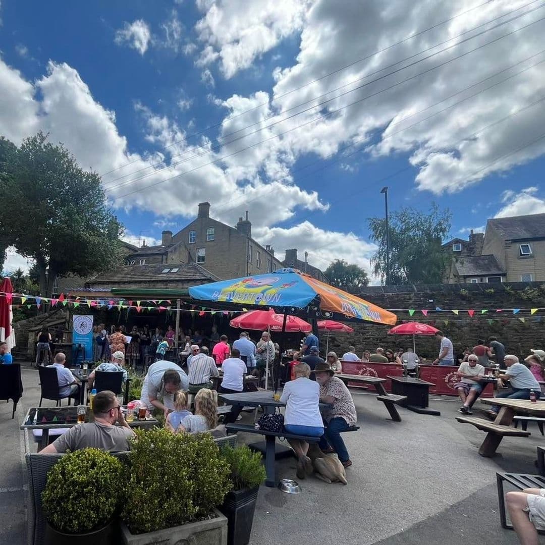 TheThornhill_OutdoorDining_SunnyDay_CommunityGathering Outdoor seating area bustling with people at The Thornhill, featuring colorful umbrellas, picnic tables, and festive bunting under a blue sky with scattered clouds.