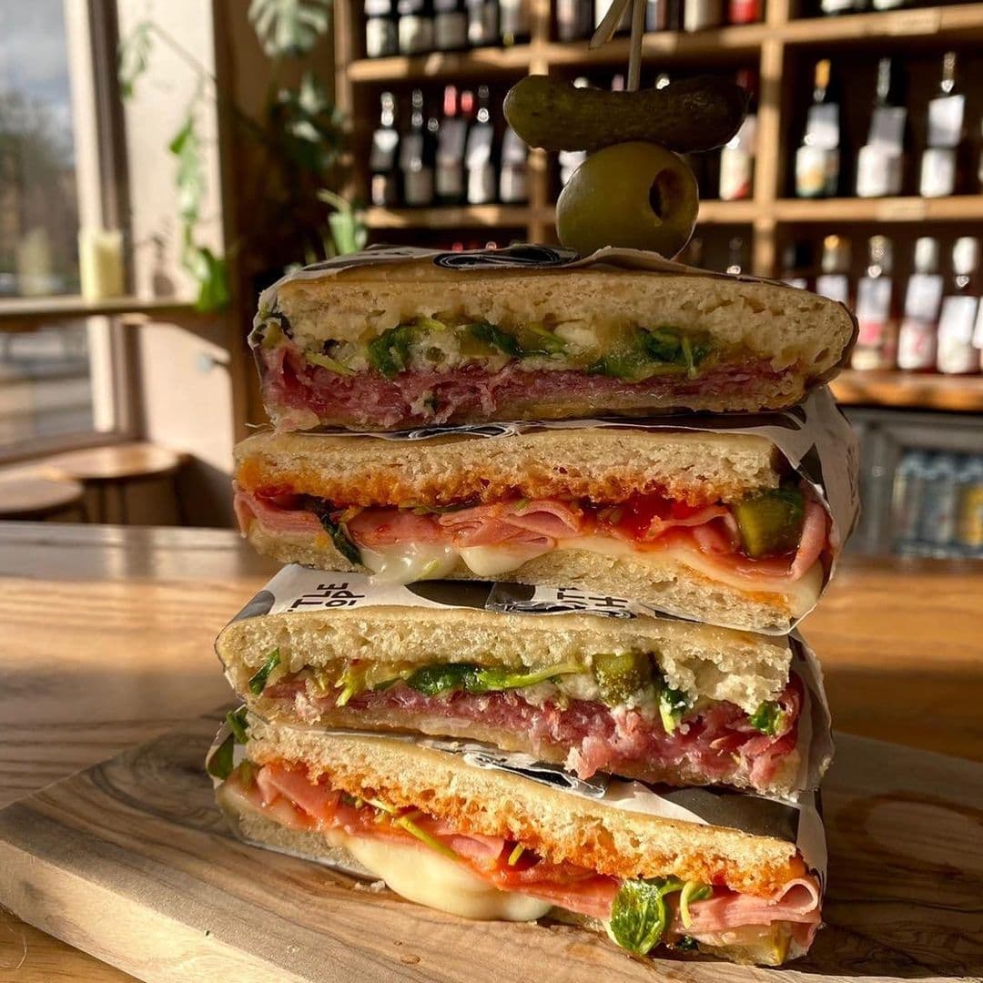 Bottle Chop Delicious Stacked Sandwiches Wooden Board Stacked deli sandwiches with various fillings on a wooden board at Bottle Chop, garnished with an olive and a pickle on top.