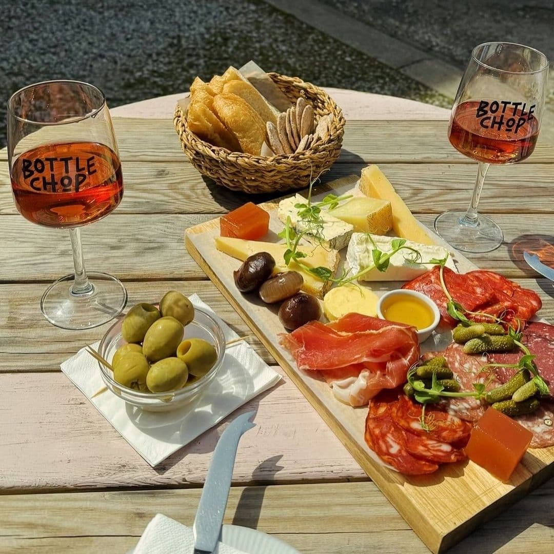 BottleChop SunnyAlfrescoDining CharcuterieCheese Wine Alt text: "A sunny outdoor dining experience at Bottle Chop featuring a glass of rose wine, a charcuterie and cheese board with assorted meats, cheeses, olives, and pickles, accompanied by a basket of fresh bread."