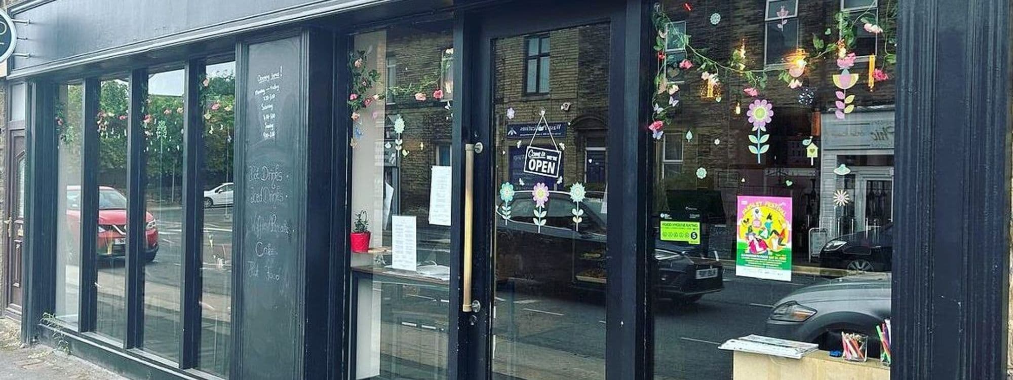 Exterior view of Cup 19 Coffee with large windows adorned with flower decorations, a sign indicating the cafe is open, and a chalkboard menu visible through the glass.