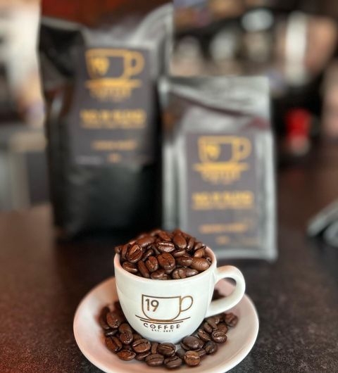 Cup 19 Coffee roasted beans cup branded bags A small white espresso cup filled with coffee beans on a saucer, branded with "Cup 19 Coffee" logo, in focus in the foreground. Blurry in the background are packaged coffee bags with the same logo.