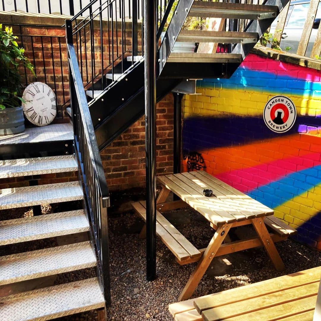 Doghouse Bar Bagel Shop Colorful Outdoor Seating Area Outdoor seating area at Doghouse Bar & Bagel Shop with a colorful rainbow-painted wall, a wooden picnic table, and metal spiral staircase.