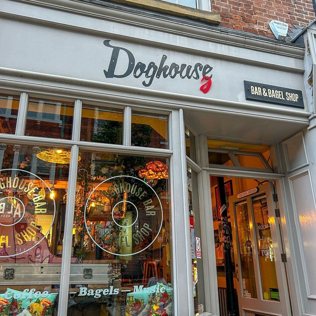Doghouse Bar Bagel Shop Cozy Storefront Evening Lights Exterior view of Doghouse Bar & Bagel Shop with signage, warm lighting, and reflections of the street on the glass windows.