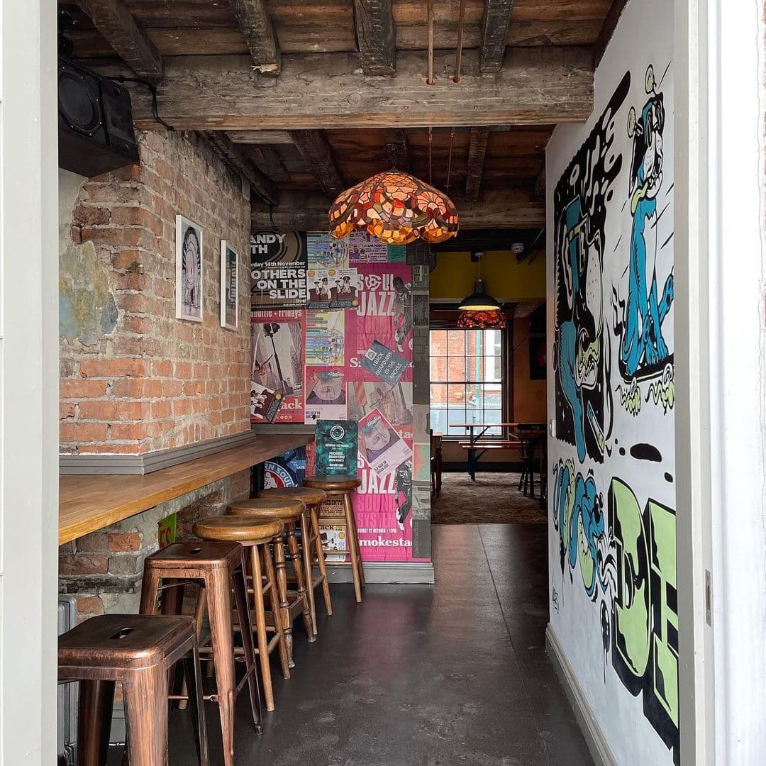 Doghouse Bar Bagel Shop Interior Exposed Brick Graffiti Decor Interior of Doghouse Bar & Bagel Shop featuring exposed brick walls, wooden bar stools, colorful posters, and unique hanging lamps.