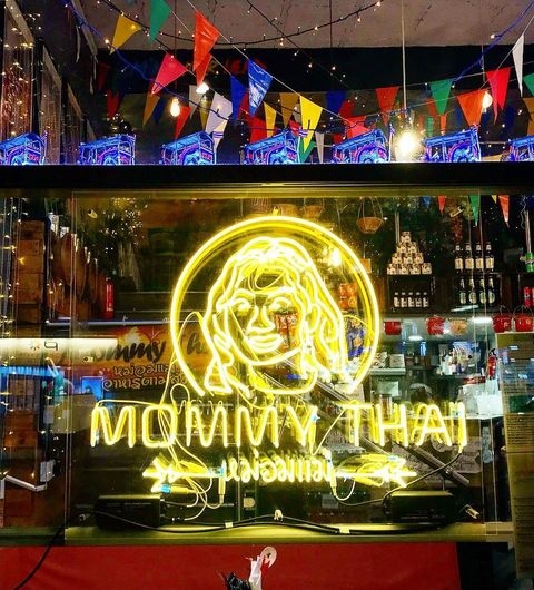 Little Mommy Thai Neon Sign Festive Decor Neon sign of Little Mommy Thai restaurant with bright yellow illumination featuring a woman's face, surrounded by colorful flags and a glimpse of the interior bar shelves.