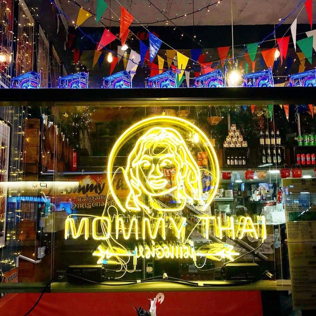 Mommy Thai Neon Sign Colorful Banners Window Display Neon sign of Mommy Thai with a smiling woman's face, surrounded by colorful flags and a glimpse of the bar interior.