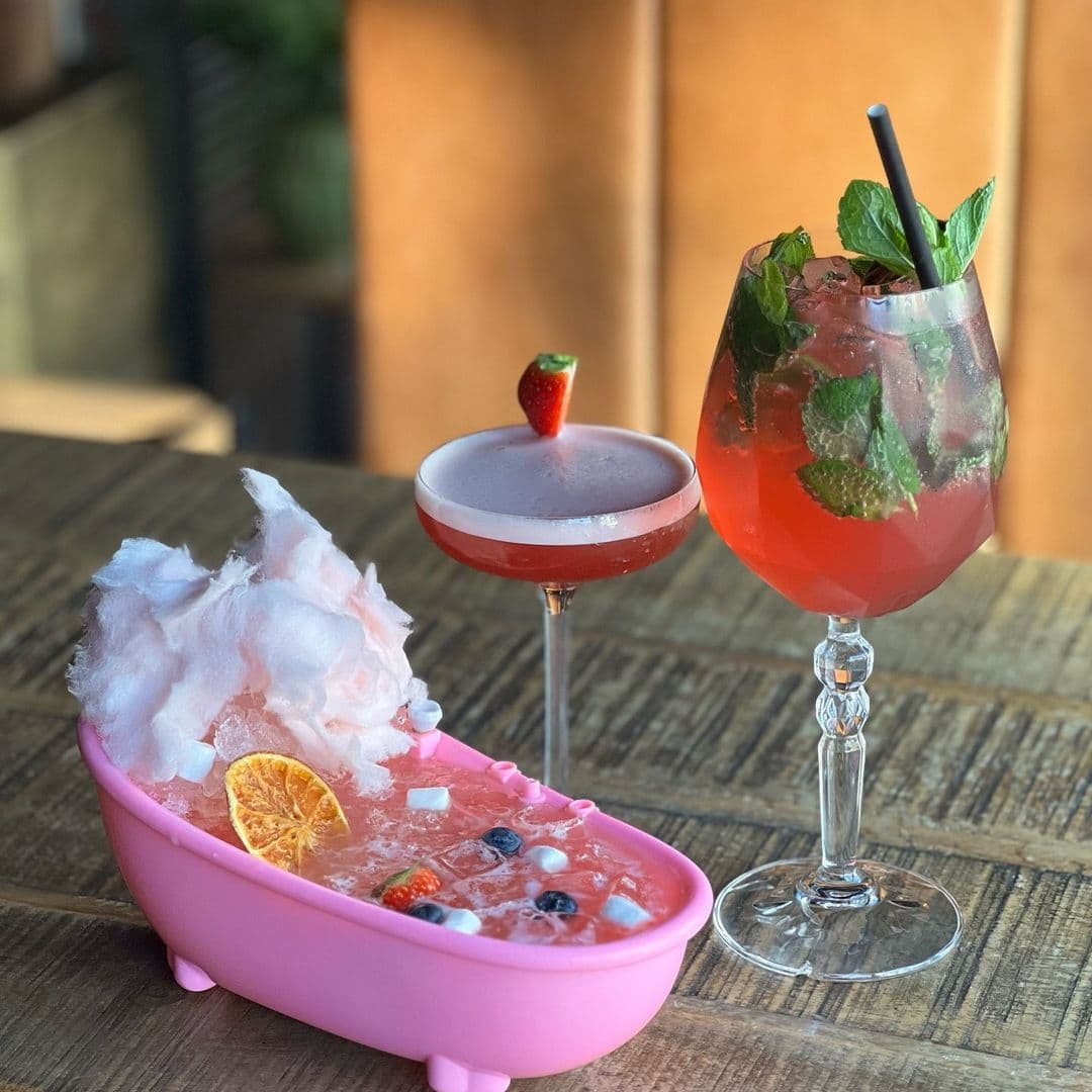 Santorini Bar and Grill Signature Cocktails Creative Presentation Creative cocktails at Santorini Bar and Grill, featuring a pink bathtub-shaped drink filled with punch and garnished with fruit, alongside a strawberry-topped martini and a minty mixed drink in a balloon glass.
