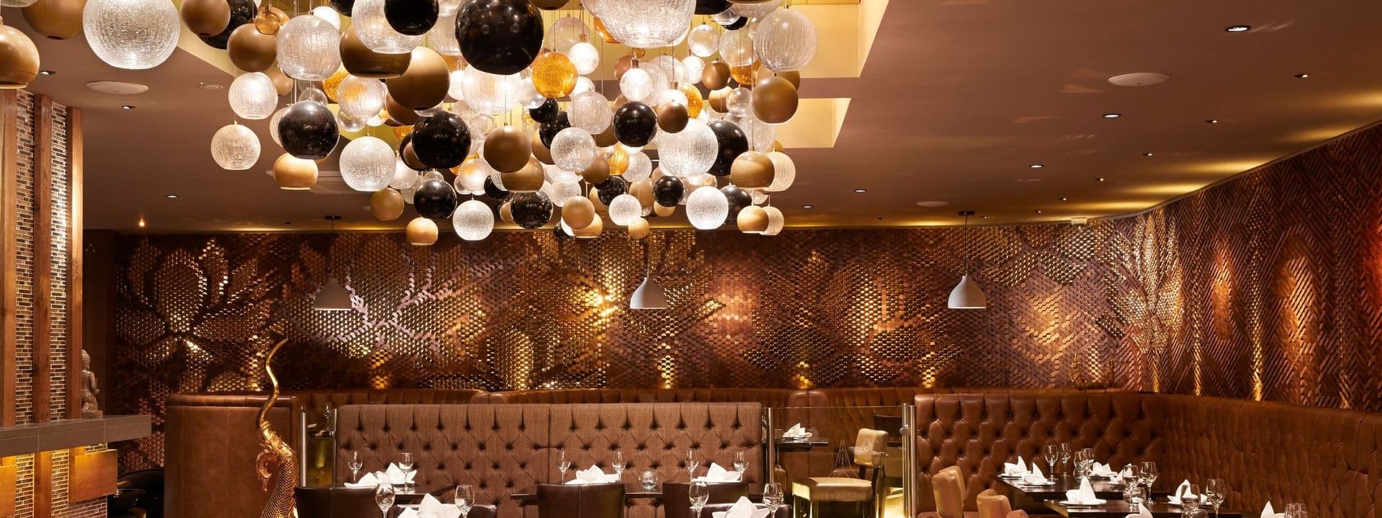 Interior of Sukhothai Restaurant – City Centre, featuring elegant dining setup with brown tufted booths, white tablecloths, and an array of spherical and textured glass pendant lights above. Decorative bronze-toned wall panels add a warm, inviting ambiance.