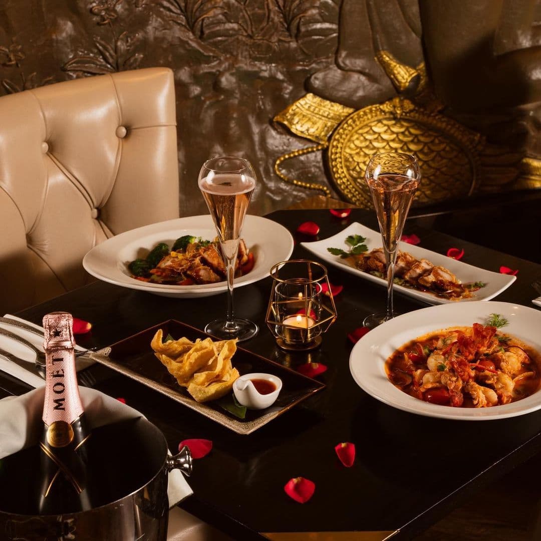 Sukhothai Restaurant City Centre Elegant Dining Experience Champagne Dinner Elegant dining setup at Sukhothai Restaurant – City Centre, featuring a bottle of champagne, two glasses, and various Thai dishes on a table adorned with rose petals and candles.