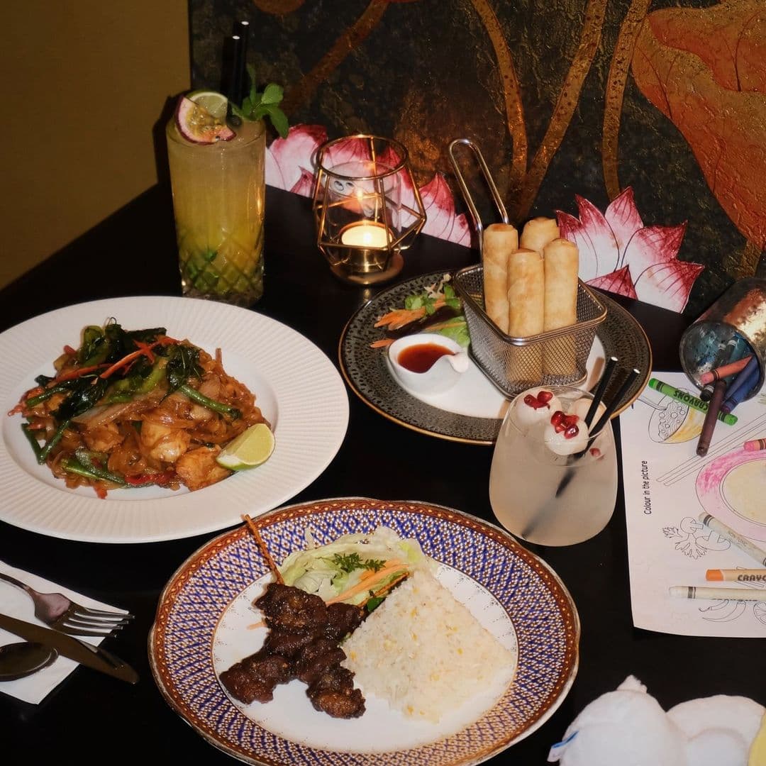 Sukhothai Restaurant City Centre Thai Cuisine Dining Ambiance A variety of Thai dishes on a table at Sukhothai Restaurant – City Centre, including stir-fried noodles with vegetables, spring rolls with dipping sauce, grilled skewered meat with rice, and a refreshing cocktail with a mint garnish. The table also features a lit candle, coloring materials for children, and a decorative backdrop.
