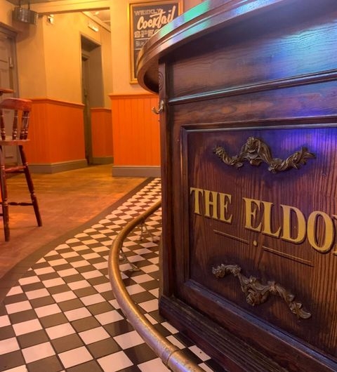 The Eldon Pub Vintage Interior Wooden Details Checkered Floor Interior of The Eldon Pub with a close-up of a wooden counter engraved with the pub's name, set against a checkered floor and warm-toned walls.