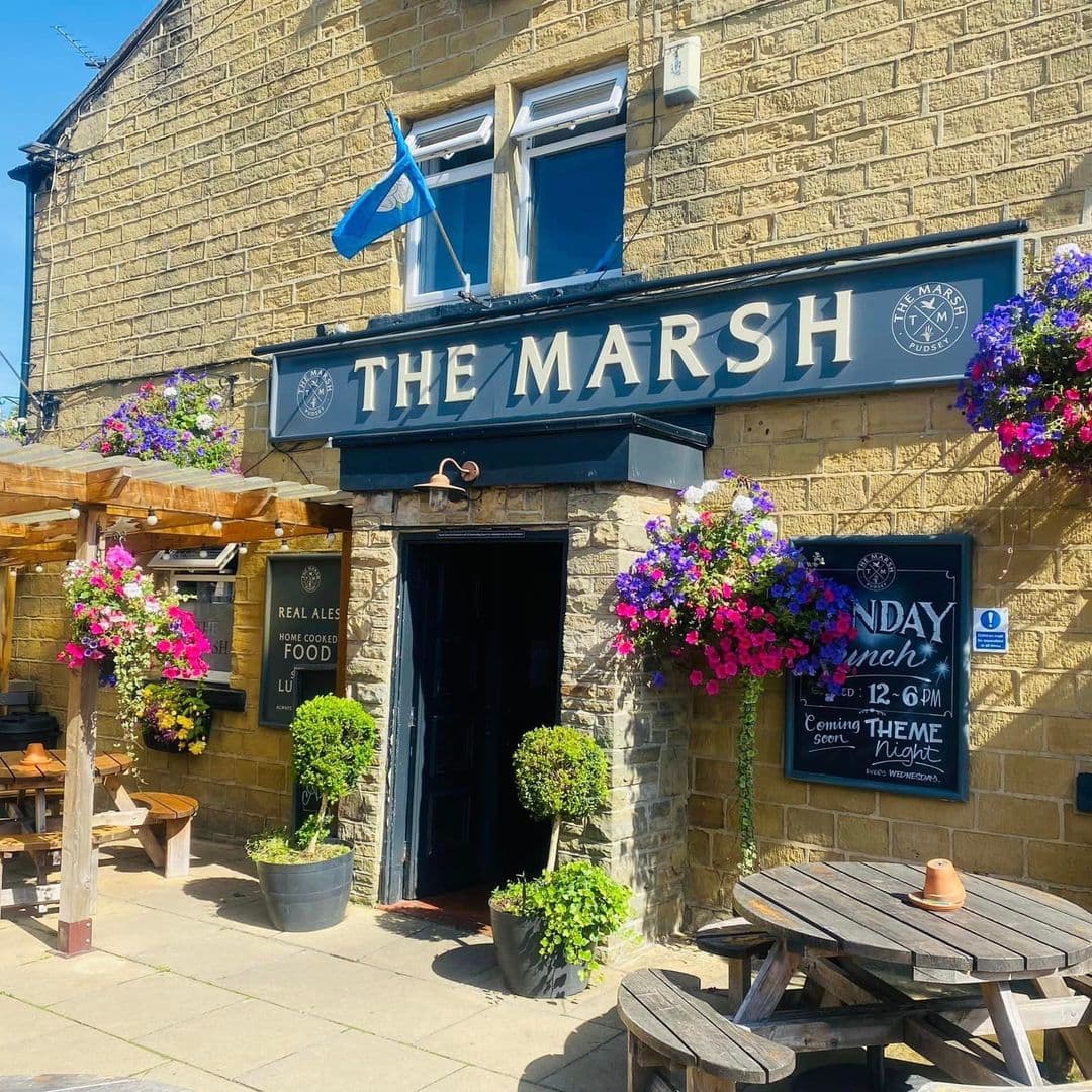 The Marsh Pudsey Traditional Pub Flower Decor Sunny Day Exterior view of The Marsh in Pudsey on a sunny day, showcasing its stone facade with a black signboard, colorful hanging flower baskets, and outdoor wooden seating.