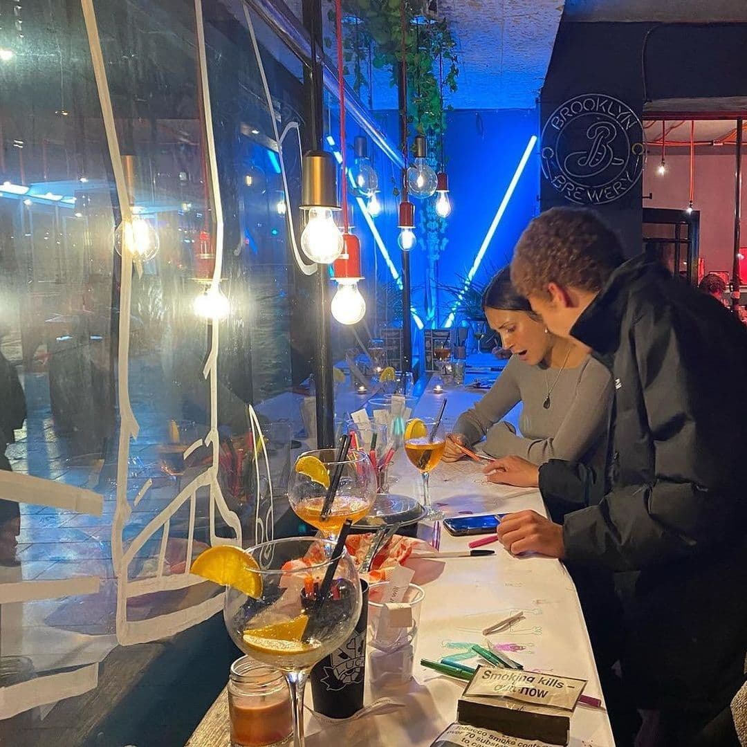 The Old Red Bus Station Creative Workshop Cocktails Nightlife People engaging in a creative activity at The Old Red Bus Station, with cocktails and art supplies on the table, and ambient lighting from hanging bulbs.