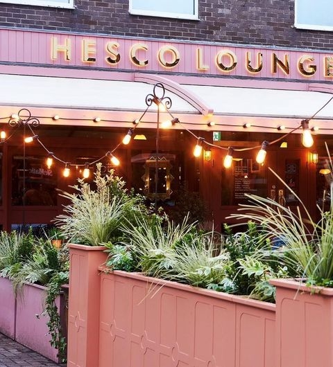HescoLounge Evening Ambiance StringLights OutdoorSeating Exterior view of Hesco Lounge with illuminated sign and string lights, featuring outdoor plants and a cozy ambiance.
