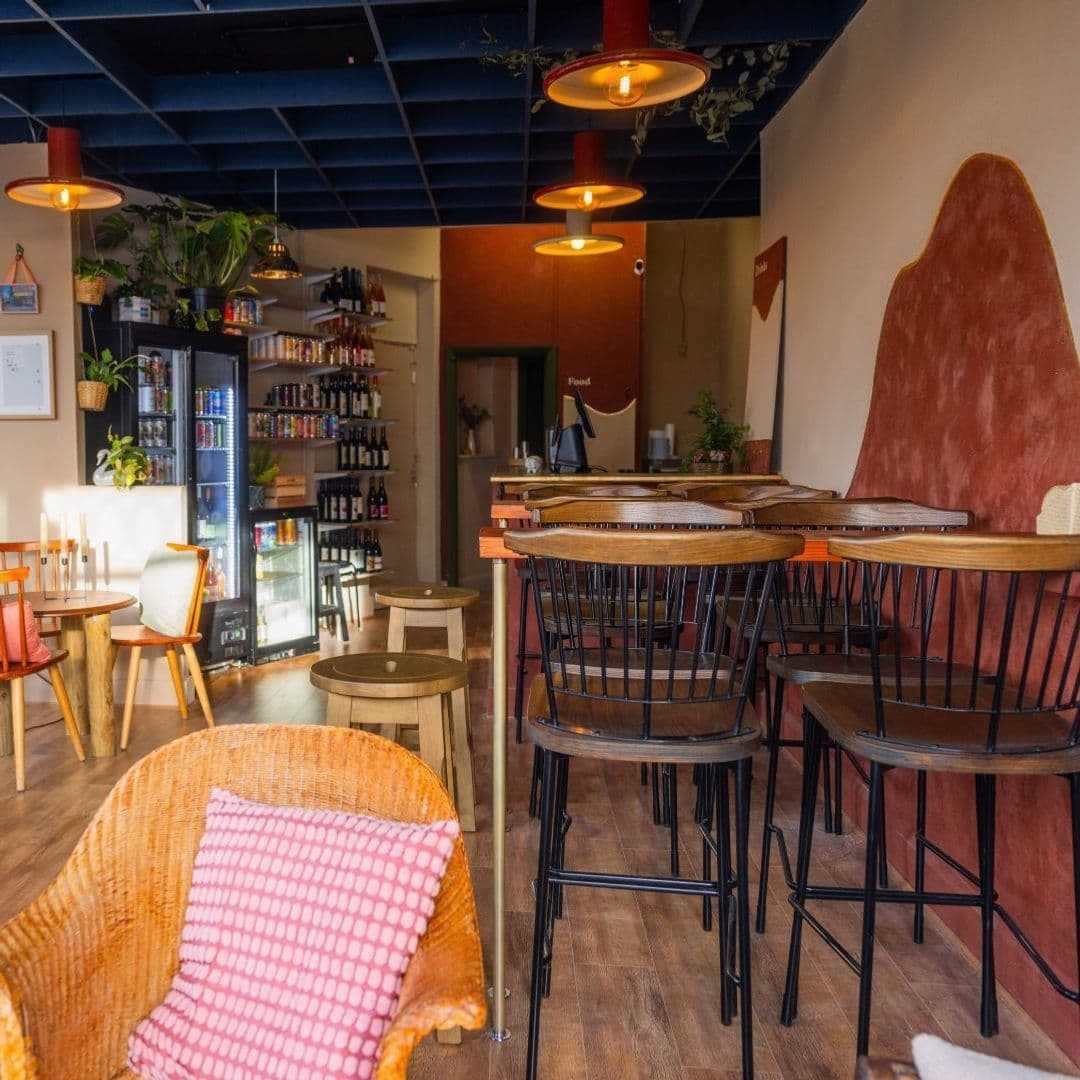 HocLoco CozyCafeInterior WarmLighting WoodenFurniture Interior of Hoc Loco featuring cozy seating, warm lighting from pendant lamps, a beverage fridge, and a welcoming ambiance with earthy tones and wooden furniture.