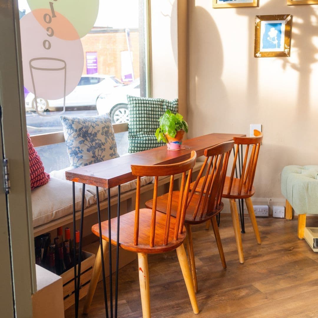 HocLoco CozyCafeInterior WoodenFurniture NaturalLight Cozy seating area at Hoc Loco with wooden chairs, a bench with cushions, and a potted plant on a table by a window with street view.