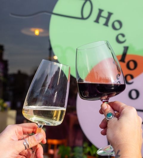 HocLoco WineToast Celebration Friends Outdoor Two hands toasting with glasses of white and red wine at Hoc Loco with the establishment's sign in the background.