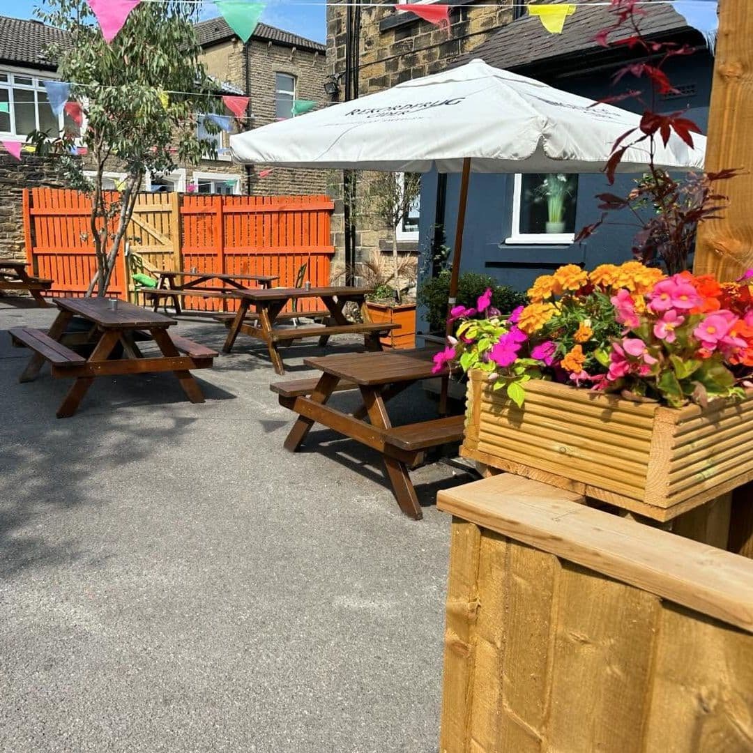 The Commercial Outdoor Seating Colorful Flowers Sunny Day Outdoor seating at The Commercial with wooden picnic tables, colorful flowers in planters, and a white umbrella on a sunny day.