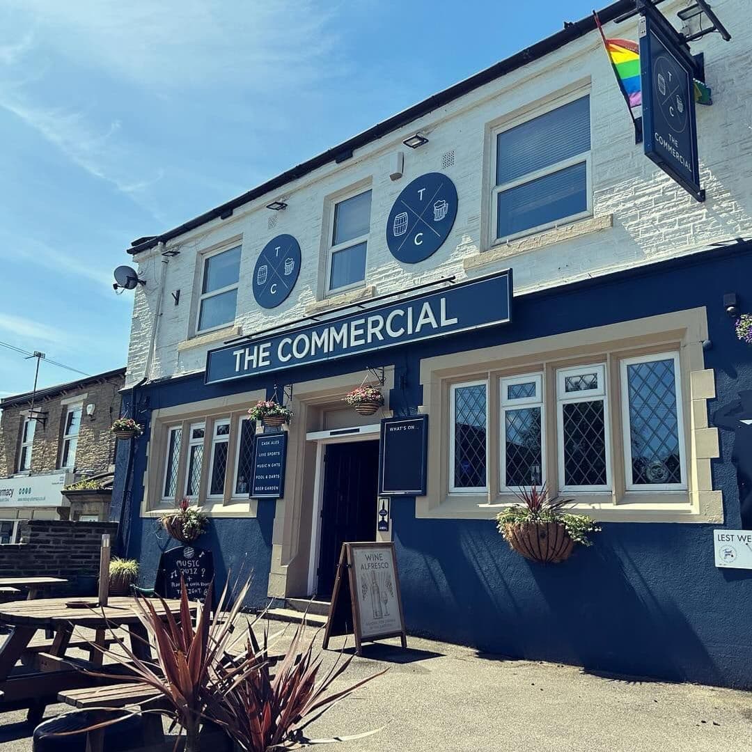 The Commercial Pub Exterior Sunny Day Flower Baskets Exterior view of The Commercial pub with blue signage, white walls, and a rainbow flag under a clear blue sky. Wooden benches are visible in the outdoor seating area.
