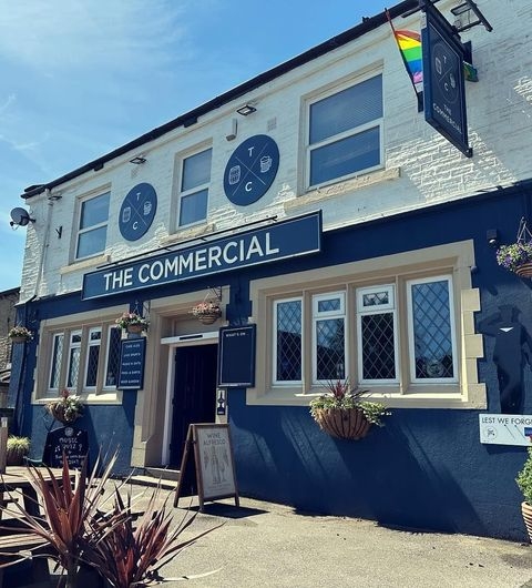 The Commercial Traditional Pub Exterior Sunny Day Exterior view of The Commercial, featuring its blue and white facade with signage, windows with flower baskets, and a clear blue sky above.