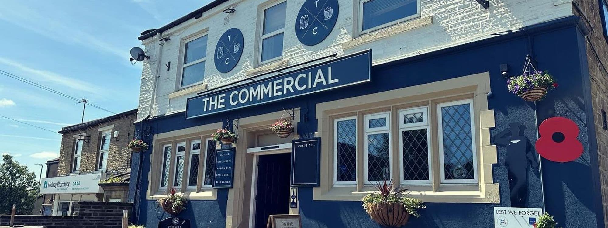 Exterior view of The Commercial, a traditional pub with a dark blue facade, white-framed windows, and hanging flower baskets, featuring a large red poppy decoration and a "Lest We Forget" sign.